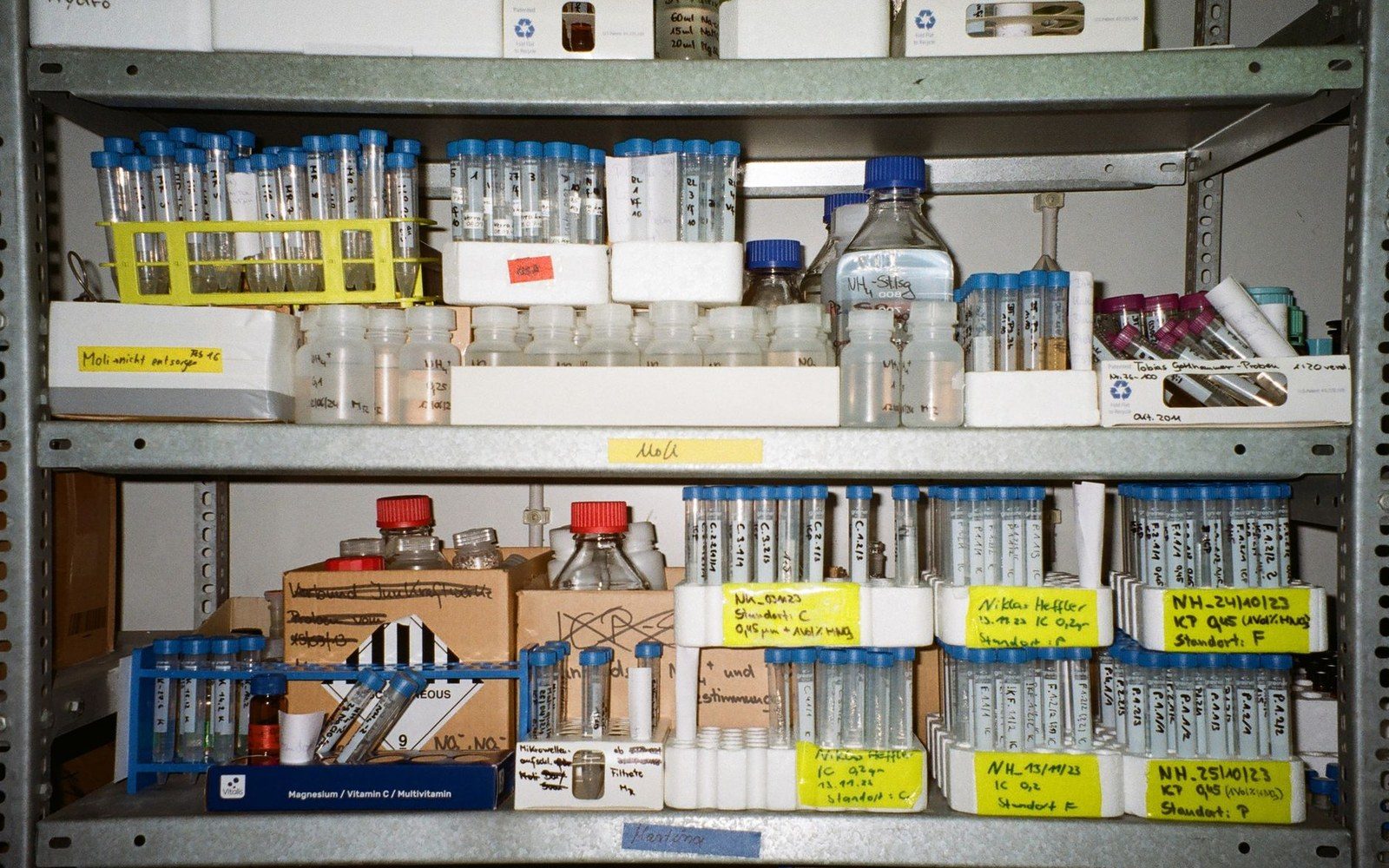 A shelf of film cans with handwritten paper labels, shot straight on, the ink marks legible on several cans in the foreground