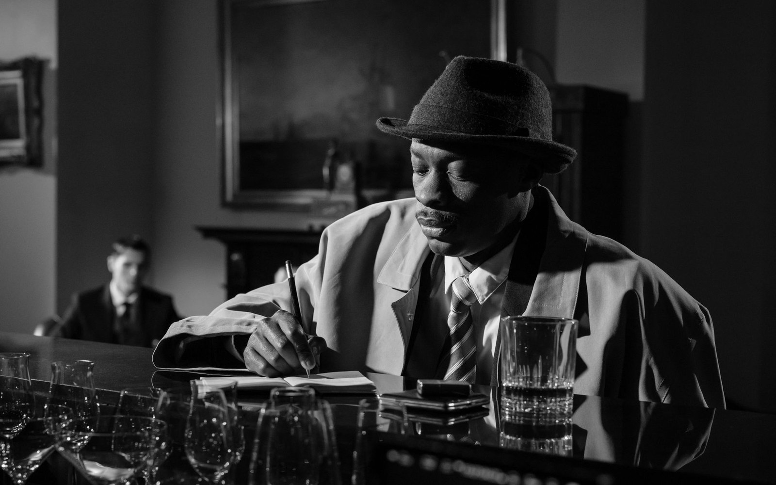 Man in trench coat and fedora writes notes at a bar counter