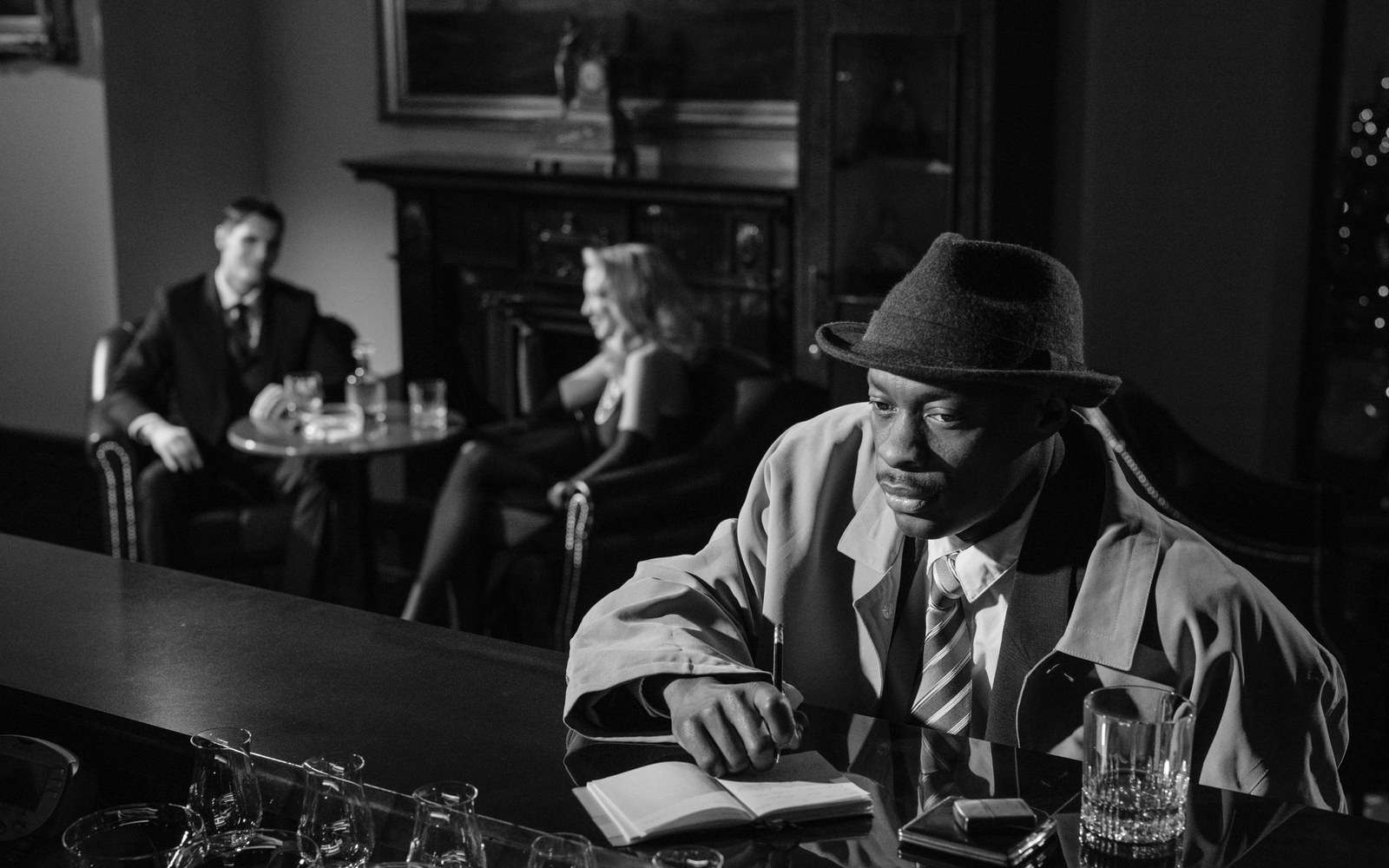 Man in trench coat and fedora writes in a notebook at a bar, couple seated behind