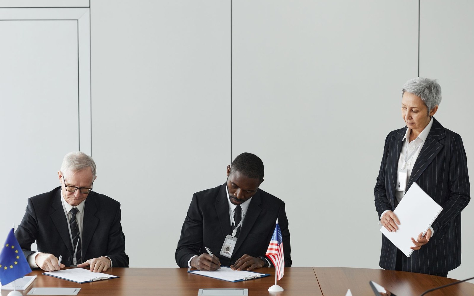 Two officials sign documents at a table with EU and US flags while a woman looks on
