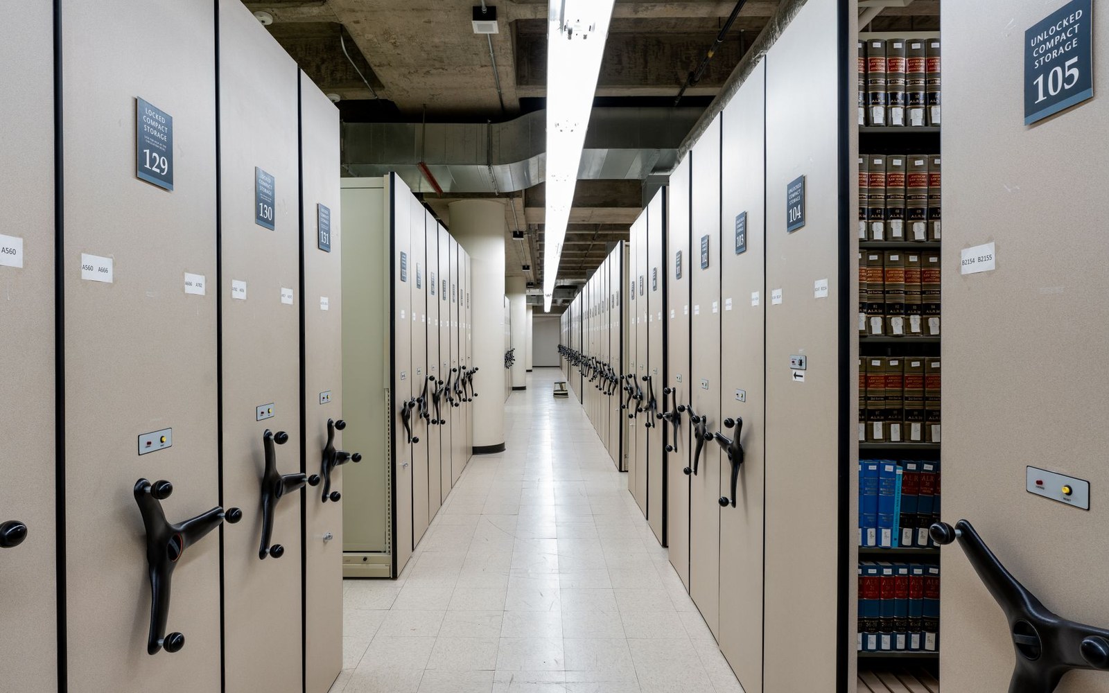 A long vault corridor with metal shelving on both sides holding labelled film cans, shot at low angle to show depth, temperature gauge visible on the wall